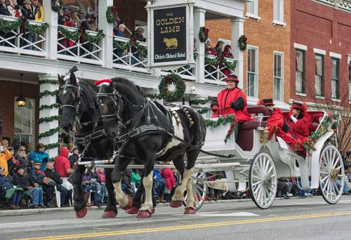A festive horse-drawn carriage with passengers in red coats, passing by a decorated hotel during a holiday parade.