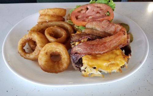 A plate with a cheeseburger topped with bacon, lettuce, tomato, pickles, and a side of onion rings.