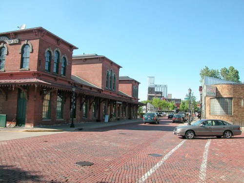 Historic brick buildings line a street with parked cars and clear blue skies in a vibrant urban setting.