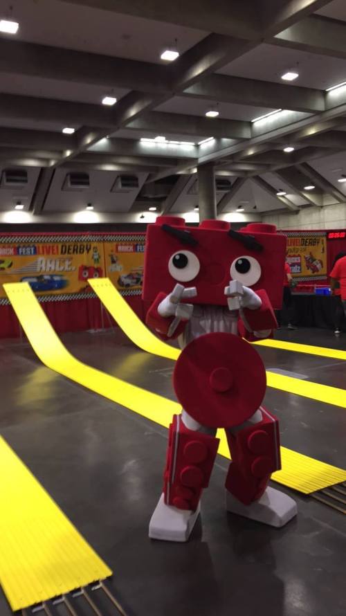 A red LEGO-inspired mascot poses with thumbs up in a convention hall, with yellow race tracks in the background.