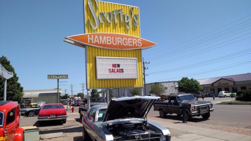 A bright yellow sign for Scotty's Hamburgers announces "New Salads," with classic cars parked nearby on a sunny day.