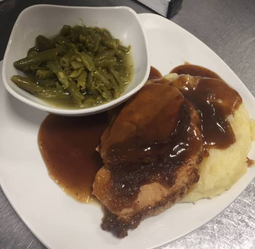 A plate of mashed potatoes topped with gravy, alongside a piece of meat and a bowl of green beans.