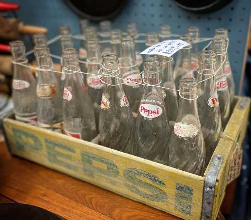 A wooden crate filled with vintage Pepsi glass bottles, some with labels, displayed on a wooden surface.