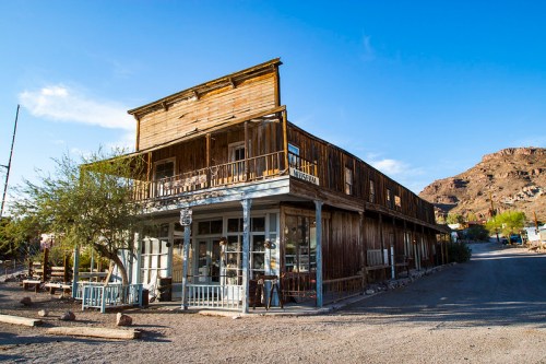 A rustic wooden building with a balcony, set against a clear blue sky and rocky landscape in a ghost town.
