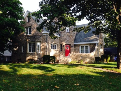 A stone house with a red door, surrounded by trees and a well-maintained lawn on a sunny day.