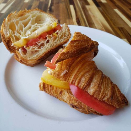 A sliced croissant filled with layers of cheese, tomato, and yellow bell pepper on a white plate.