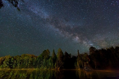 A starry night sky with the Milky Way over a calm lake, surrounded by trees reflecting in the water.