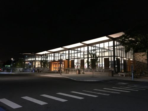 A modern building with large glass windows, illuminated at night, surrounded by trees and a parking lot.