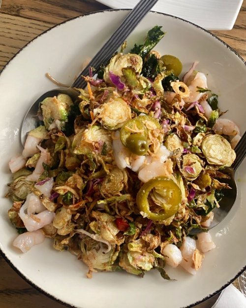 A colorful salad with Brussels sprouts, shrimp, red onion, and jalapeños, served in a bowl with two spoons.