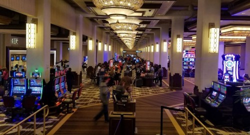 A bustling casino interior with rows of slot machines and gaming tables, illuminated by chandeliers and colorful lights.