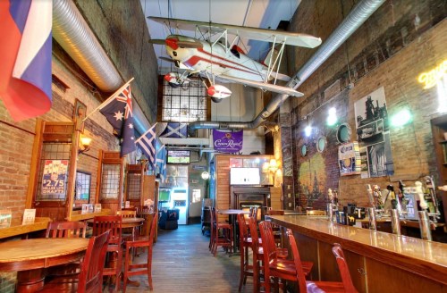A rustic bar interior with wooden chairs, flags hanging, and a model airplane suspended from the ceiling.