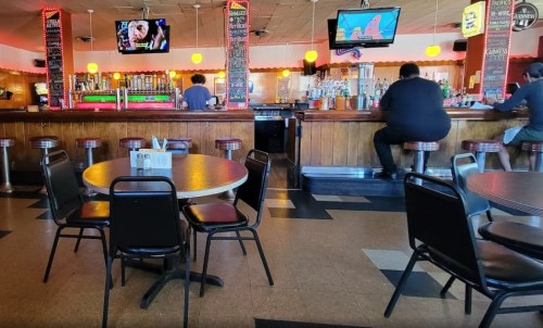 A view of a bar with empty tables, colorful lights, and two patrons sitting at the counter. TVs display images above.