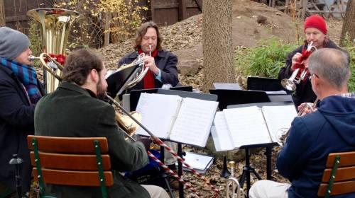 A small brass ensemble performs outdoors, surrounded by trees and autumn leaves, with sheet music on stands.