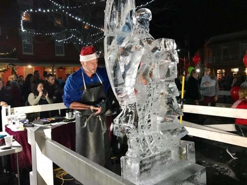 A man in a Santa hat carves an ice sculpture at a festive night event, surrounded by an audience and holiday lights.