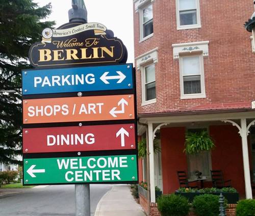 Signpost in Berlin with directions for parking, shops/art, dining, and welcome center. Red brick building in background.