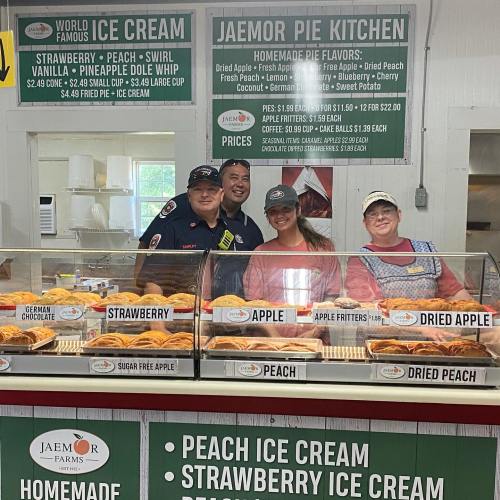 A group of four people stands behind a counter displaying various pies and ice cream flavors at a farm market.
