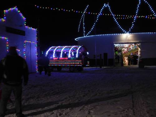 A festive scene at night with a brightly lit wagon and buildings adorned with colorful holiday lights. Snow covers the ground.