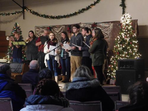 A group of singers performs on stage in front of a decorated Christmas tree, with an audience seated in front.
