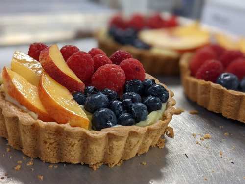 Fresh fruit tarts topped with raspberries, blueberries, and peach slices on a silver surface.