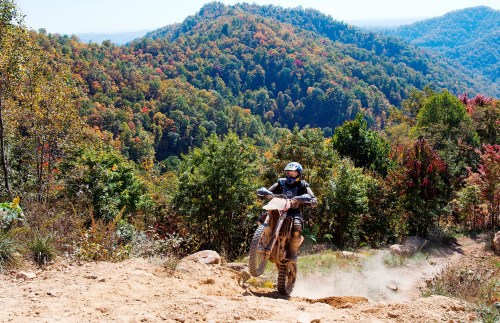A motorcyclist performs a wheelie on a dirt bike in a scenic, colorful mountain landscape.
