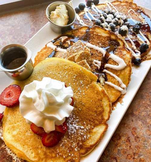 A plate of fluffy pancakes topped with strawberries, blueberries, whipped cream, and drizzled with syrup and icing.