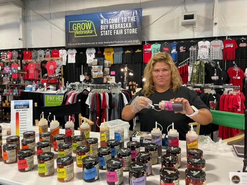 A vendor at the Nebraska State Fair Store displays jars of sauces while preparing samples for visitors.