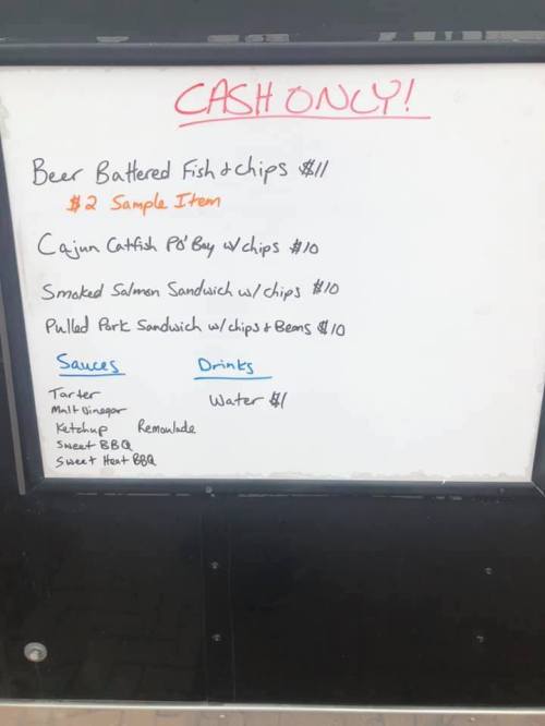 Menu board listing cash-only food items: fish & chips, catfish, salmon sandwich, pulled pork, sauces, and drinks.