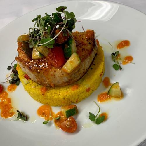 A beautifully plated dish featuring seared fish atop yellow couscous, garnished with microgreens and colorful sauces.
