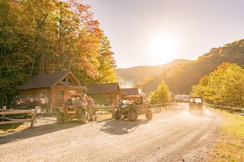 Two people work on machinery near rustic cabins, surrounded by colorful autumn trees and a setting sun in the background.