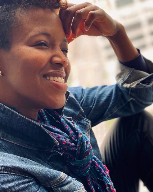 A smiling person with curly hair, wearing a denim jacket and a colorful scarf, resting their chin on their hand.