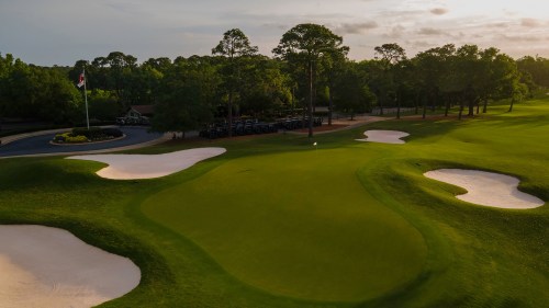 Aerial view of a golf course with green fairways, sand traps, and a parking area in the background under a cloudy sky.