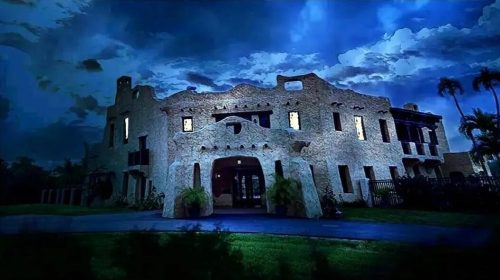 A historic stone building illuminated at night, surrounded by dark clouds and palm trees.