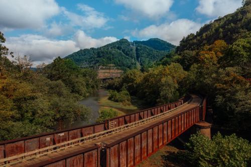 A rusted bridge spans a river, surrounded by lush green hills and a blue sky with scattered clouds.