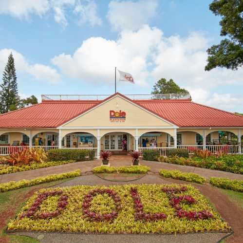 Dole Plantation building with a red roof, surrounded by colorful landscaping and a blue sky with clouds.