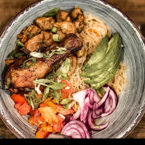 A bowl of noodles topped with grilled chicken, avocado, diced tomatoes, and red onion, garnished with green onions.