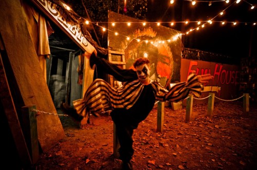 A clown in striped costume poses playfully in a dimly lit carnival setting with string lights and colorful backdrops.