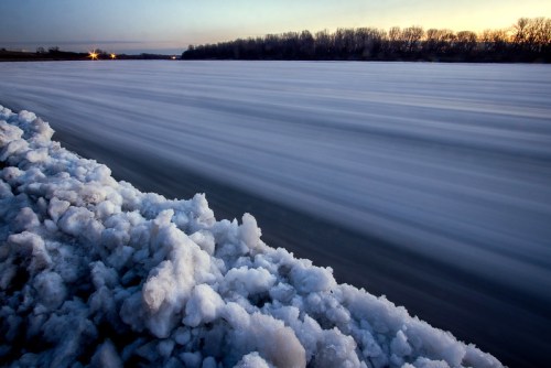 A frozen river with snow along the bank, reflecting soft light from the sunset in the background.