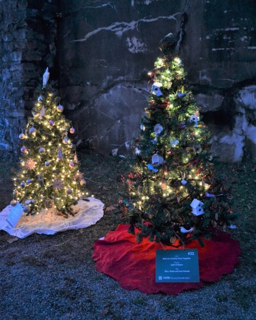 Two decorated Christmas trees with lights, set against a stone wall, on a grassy area with red and white tree skirts.
