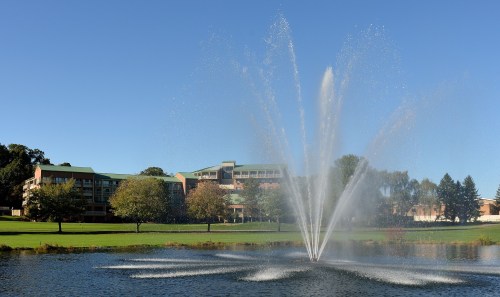 A fountain sprays water in a pond, with green lawns and buildings in the background under a clear blue sky.