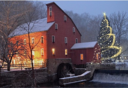 A red mill illuminated at night, with a snowy landscape and a sparkling Christmas tree beside a flowing river.