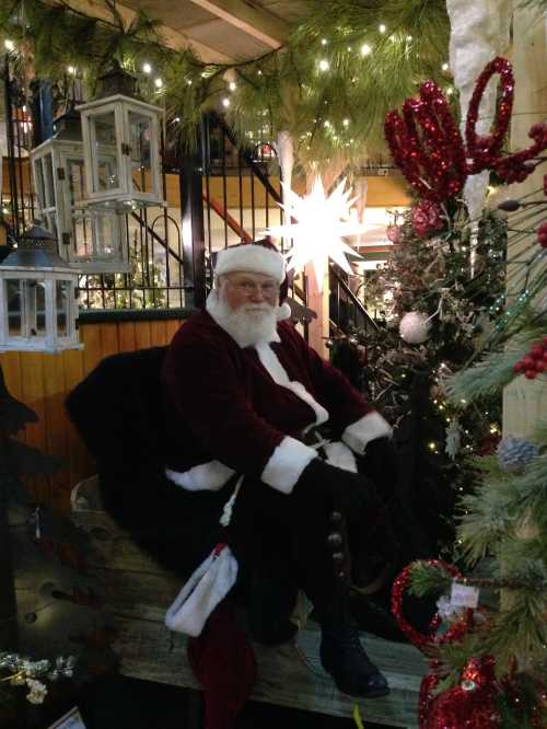 A jolly Santa Claus sits in a festive setting, surrounded by Christmas decorations and a tree.