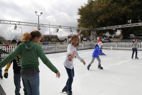 A group of children ice skating on an outdoor rink, with festive decorations in the background.
