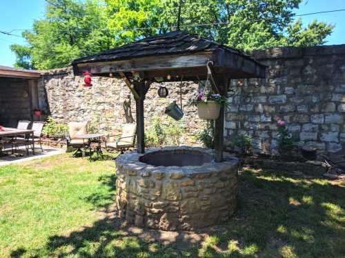 A rustic stone well with a wooden roof, surrounded by a garden and patio furniture under a clear blue sky.