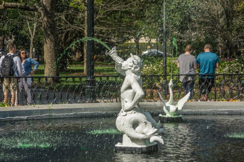 A fountain featuring a mermaid statue spouting water, with people enjoying the park in the background.