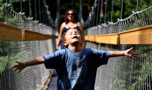A child joyfully stretches his arms on a suspension bridge, with a woman smiling in the background.
