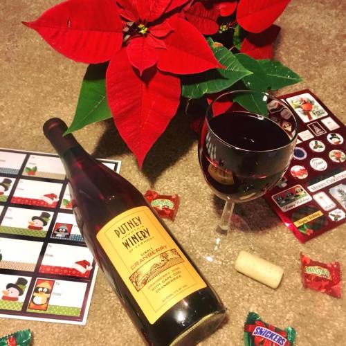 A bottle of cranberry wine, a glass of wine, and festive candies beside a poinsettia plant on a carpeted surface.