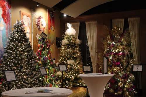 A festive display of decorated Christmas trees in a warmly lit room, with tables and colorful artwork in the background.