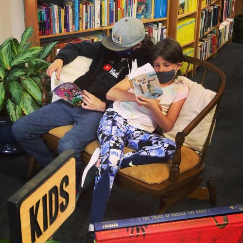 Two children reading books on a cozy couch in a library, surrounded by colorful shelves filled with books.