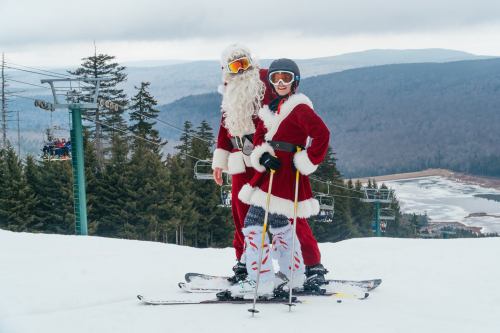 Two people in Santa costumes skiing on a snowy slope, with trees and mountains in the background.