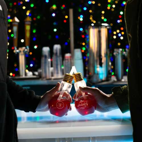 Two hands clink lightbulb-shaped glasses filled with red liquid, with a festive, colorful background.